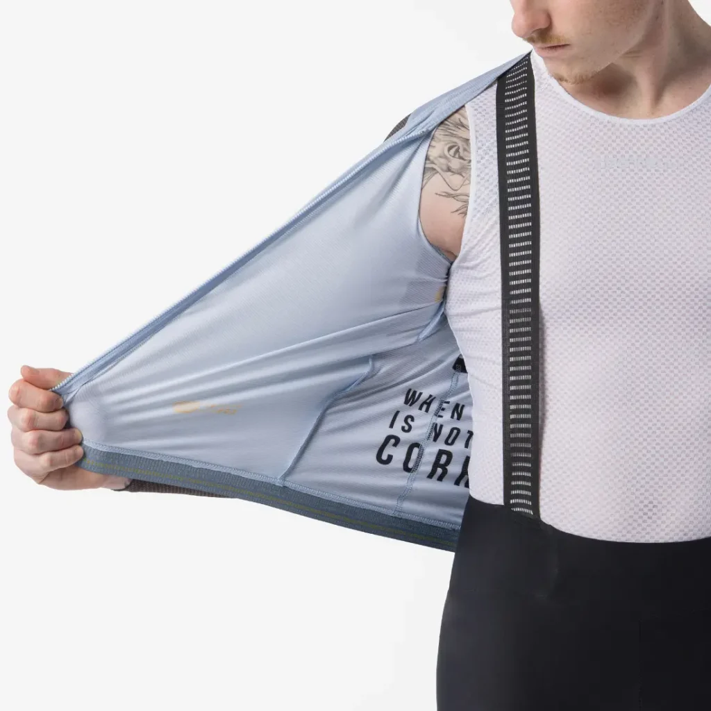 Una persona vestida con ropa de ciclismo sostiene abierto un chaleco Castelli Transition de color azul claro, dejando a la vista una camiseta interior de malla blanca y tirantes negros, perfectos para la temporada de manga larga o para combinar con tu maillot Corretto LS favorito.
