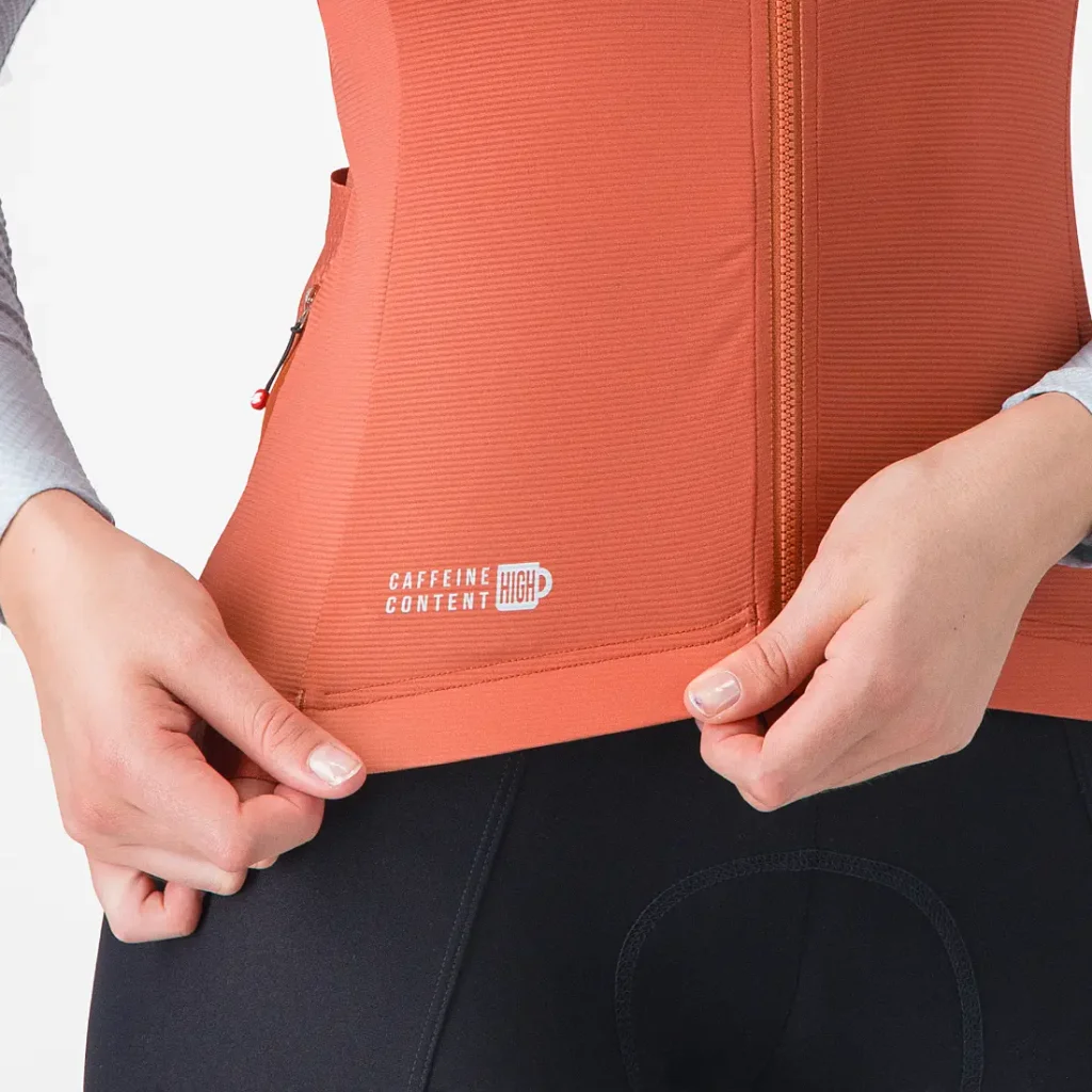 Persona que lleva un chaleco deportivo naranja con cremallera sobre una camiseta Corretto de manga larga con la inscripción «alto contenido en cafeína» cerca del dobladillo, tirando del borde inferior; solo se ven el torso y las manos.
