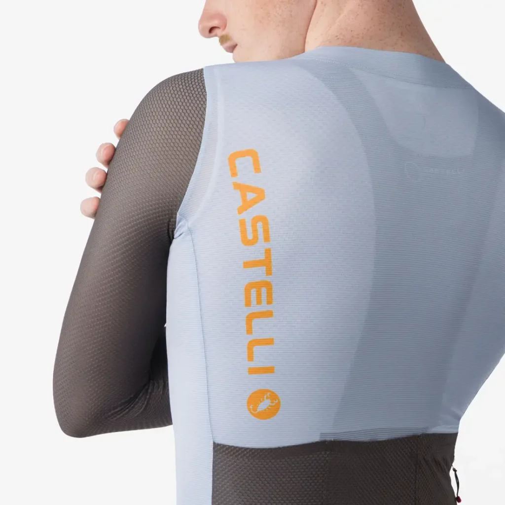 Persona con un maillot de ciclismo de manga larga azul claro y negro, con la inscripción «CASTELLI» y el logotipo de un escorpión impresos en naranja en la espalda, vista desde atrás con los brazos cruzados.