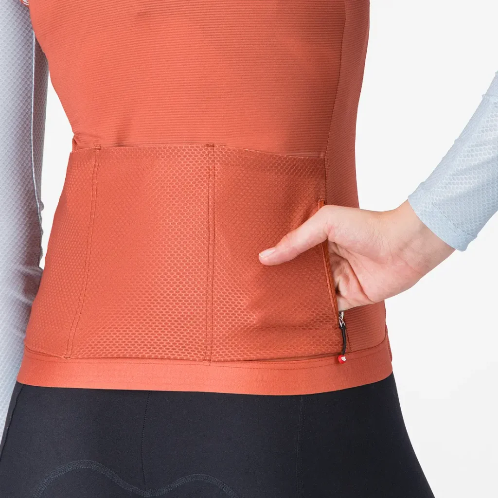 Persona que lleva una camiseta deportiva naranja «Castelli Transition» de manga larga y mallas negras, con la mano derecha metida en un bolsillo trasero con cremallera. Solo se ven el torso y la mano.