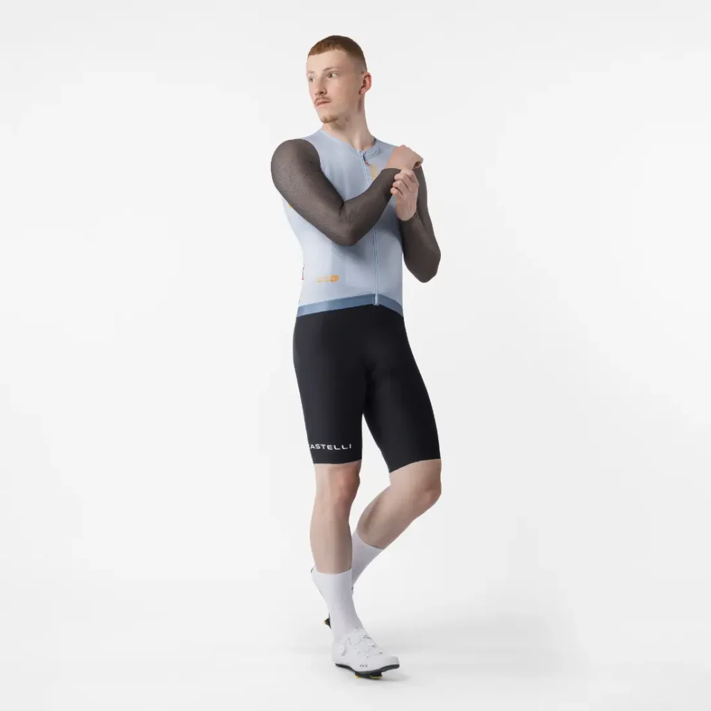 Una persona posa sobre un fondo blanco con un maillot de ciclismo sin mangas, pantalones cortos negros, calcetines blancos y zapatillas de ciclismo, mientras se ajusta el brazo derecho como si se estuviera preparando para ponerse encima el maillot Corretto LS y así ganar en comodidad durante las salidas con manga larga.