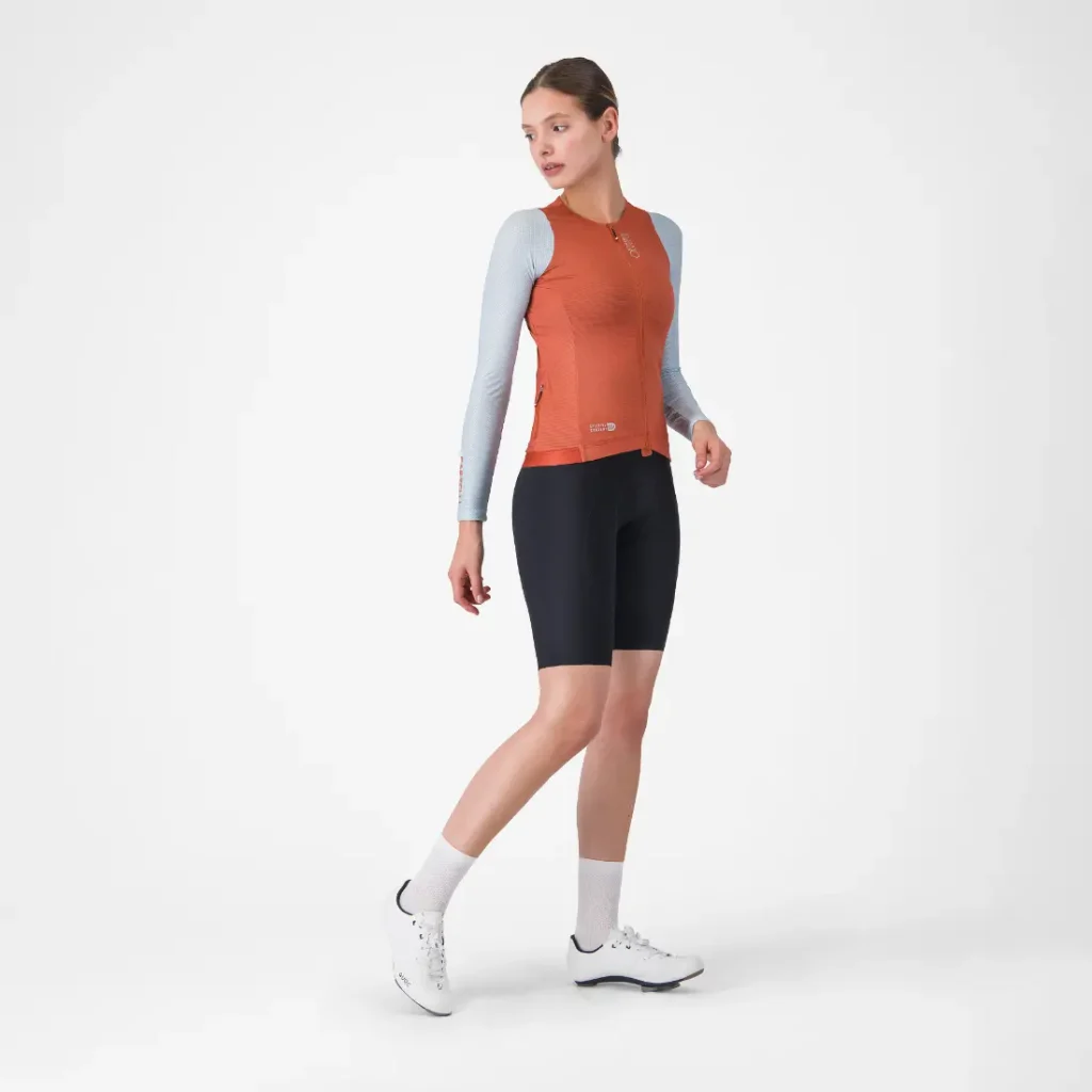 Mujer que lleva un maillot Corretto LS de manga larga en naranja vivo, culotes de ciclismo negros, calcetines blancos y zapatillas deportivas blancas, de pie frente a un fondo blanco liso.