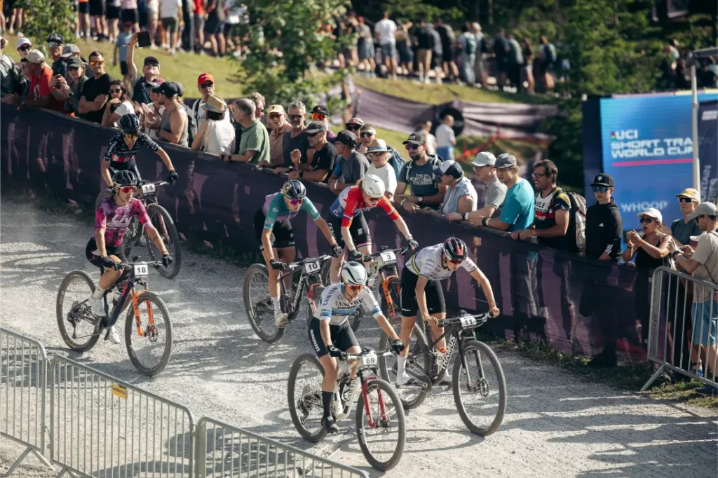 Seis ciclistas compiten en una carrera de bicicleta de montaña con ruedas de 32, mientras los espectadores observan desde detrás de las vallas. Al fondo se ve un cartel de la Copa del Mundo de Short Track de la UCI, lo que pone de relieve la revolución necesaria que se está produciendo en el sector.