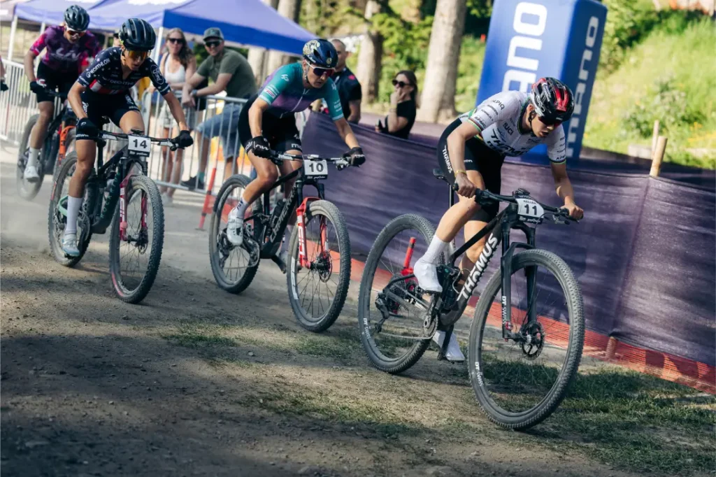 Cuatro ciclistas compiten por un sendero de tierra con ruedas de 32 durante una prueba de ciclismo de montaña, poniendo de manifiesto la revolución de la industria, mientras se ven espectadores y pancartas al fondo.