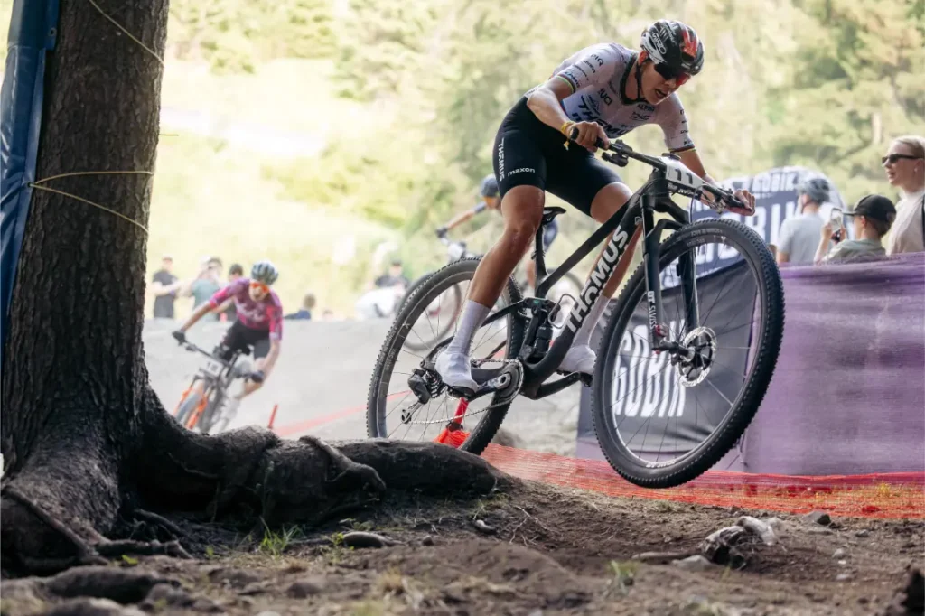Dos ciclistas compiten por un sendero de tierra que atraviesa una zona boscosa; el ciclista que va en cabeza, sorteando con destreza las raíces con sus ruedas de 32, llama la atención de los espectadores que observan desde detrás de una barrera.