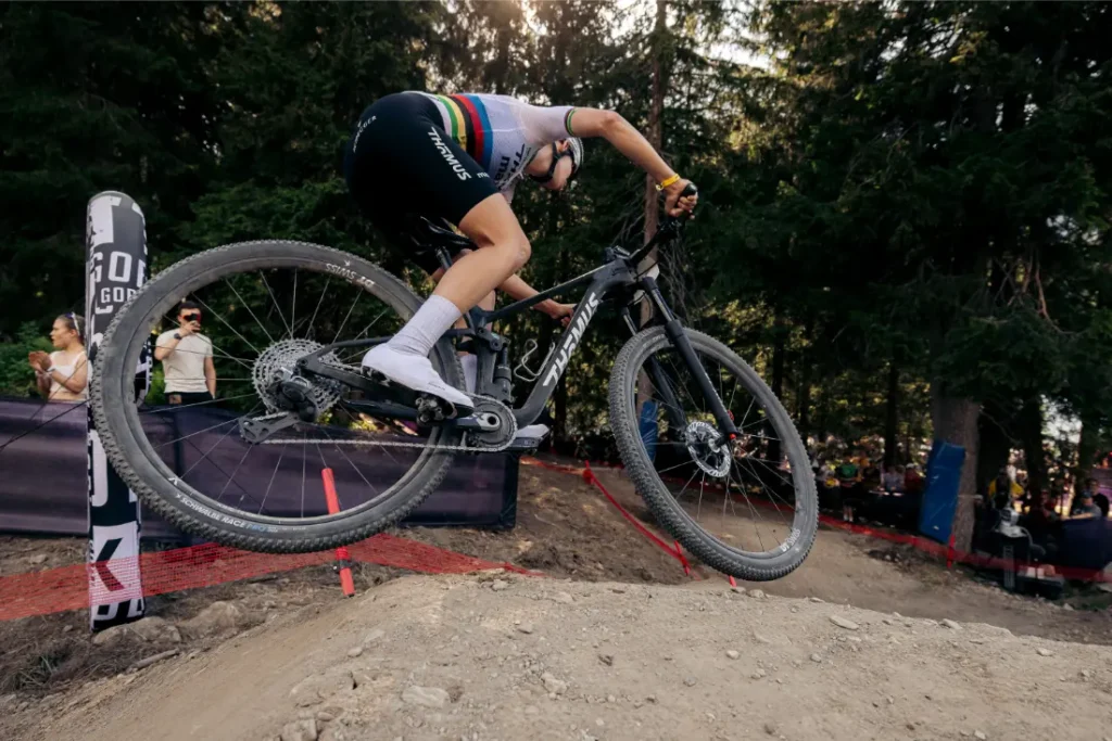 Un ciclista, con casco y equipación de competición, recorre en una bicicleta de montaña equipada con ruedas de 32 un montículo de tierra en un sendero forestal durante una prueba deportiva, mostrando las últimas tendencias de la industria del ciclismo.