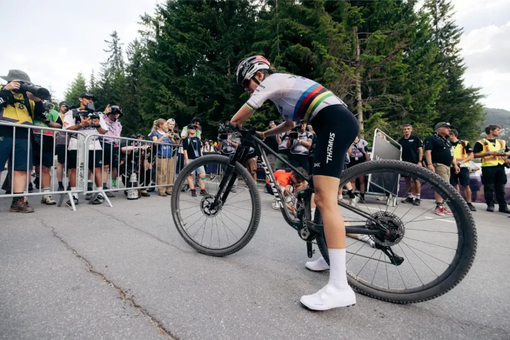 Un ciclista que lleva un maillot con rayas de colores del arcoíris está de pie junto a su bicicleta, equipada con ruedas de 32”, cerca de un grupo de fotógrafos y espectadores en un evento al aire libre.