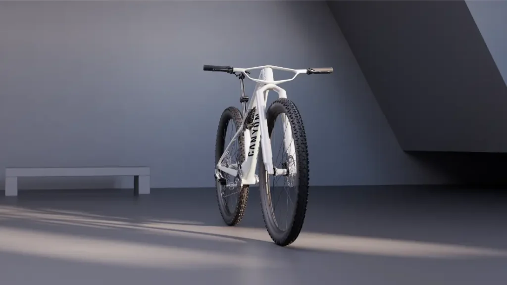 Una bicicleta de montaña Canyon blanca con ruedas de 32 está colocada en el interior, sobre un suelo gris, con la luz entrando por la izquierda y una pared gris y un banco al fondo.