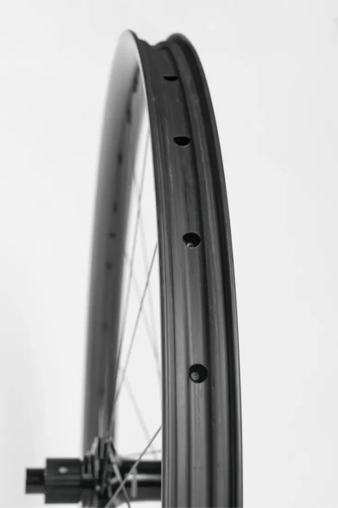 Primer plano de la llanta de una bicicleta negra en el que se aprecian los orificios de los radios y parte del eje, sobre un fondo blanco liso.