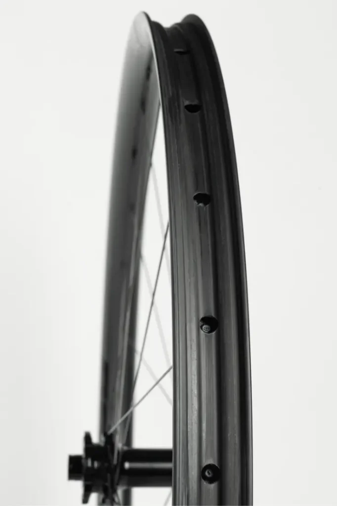 Primer plano de una llanta de bicicleta negra con los agujeros de los radios visibles, vista de perfil sobre un fondo blanco liso.