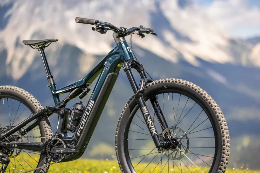 Una bicicleta de montaña eléctrica FOCUS de color negro, con ruedas grandes, está aparcada sobre la hierba, con las montañas y un fondo natural difuminado.