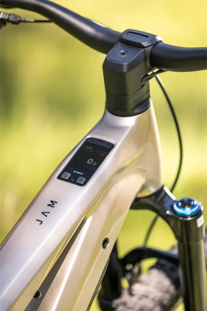 Primer plano del cuadro metálico de una bicicleta eléctrica de montaña JAM e-MTB 2027, con una pantalla digital integrada en el tubo superior, manillar negro y suspensión delantera visible, sobre un fondo difuminado al aire libre.