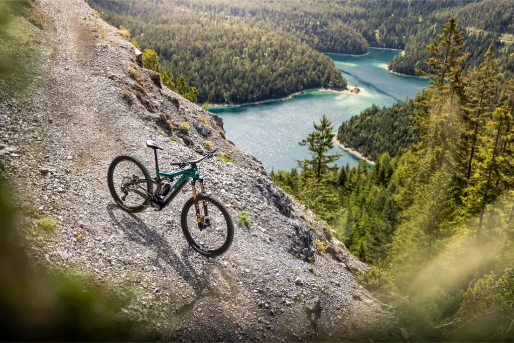 Una bicicleta de montaña FOCUS e-MTB 2027 descansa sobre una ladera rocosa con vistas a un valle boscoso y a un sinuoso lago azul que se extiende más abajo.