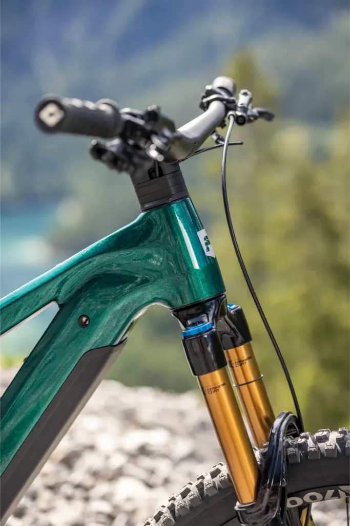 Primer plano del manillar y la horquilla de suspensión delantera de una bicicleta de montaña eléctrica Gama FOCUS e-MTB 2027 de color verde, fotografiada al aire libre con un fondo natural desenfocado.