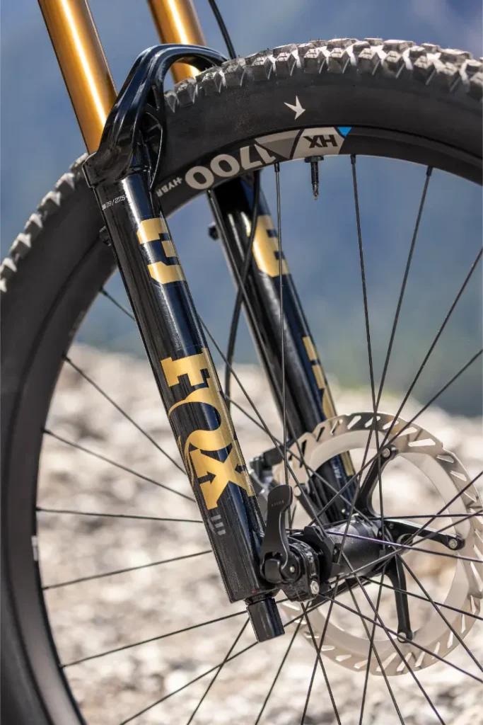 Primer plano de una horquilla de suspensión delantera de una bicicleta de montaña con el logotipo «FOX» en dorado y negro, un disco de freno y un fondo rocoso, que refleja a la perfección el rendimiento resistente que cabría esperar de una e-MTB de FOCUS como la JAM o la SAM.