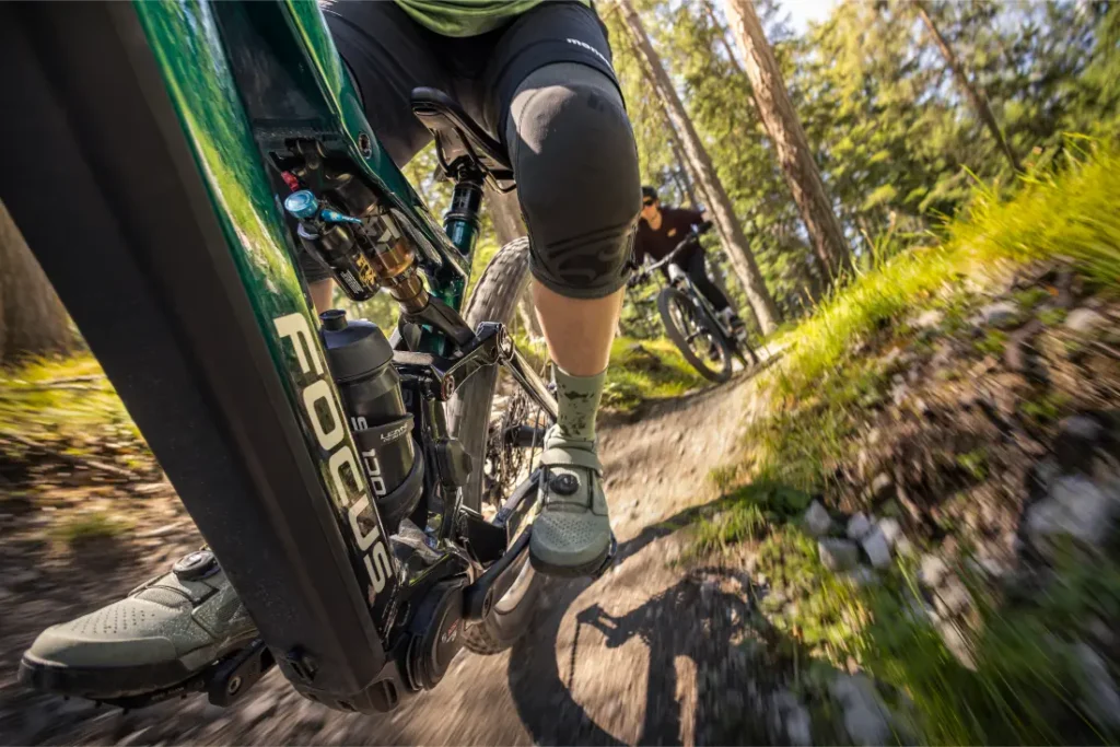 Primer plano de dos ciclistas montados en bicicletas de montaña de la gama Gama FOCUS e-MTB 2027 en un sendero forestal, con especial atención al cuadro de la bicicleta, las piernas y el movimiento dinámico.