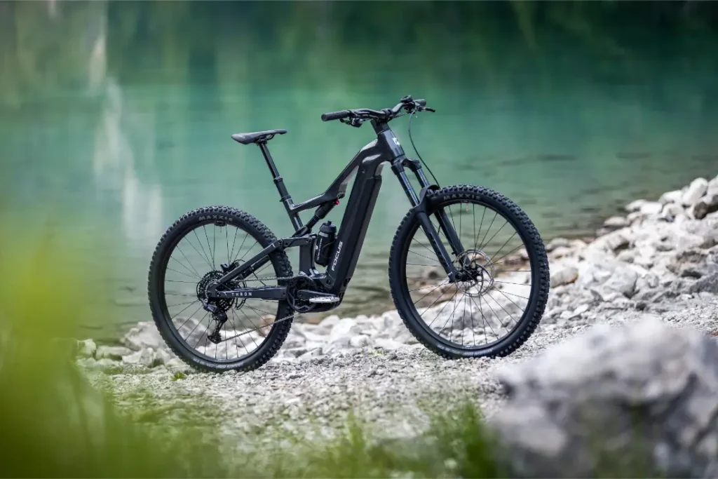 Una bicicleta eléctrica de montaña FOCUS 2027 de color negro, con neumáticos gruesos, está aparcada en un sendero rocoso junto a una masa de agua de color turquesa, con vegetación difuminada en primer plano.