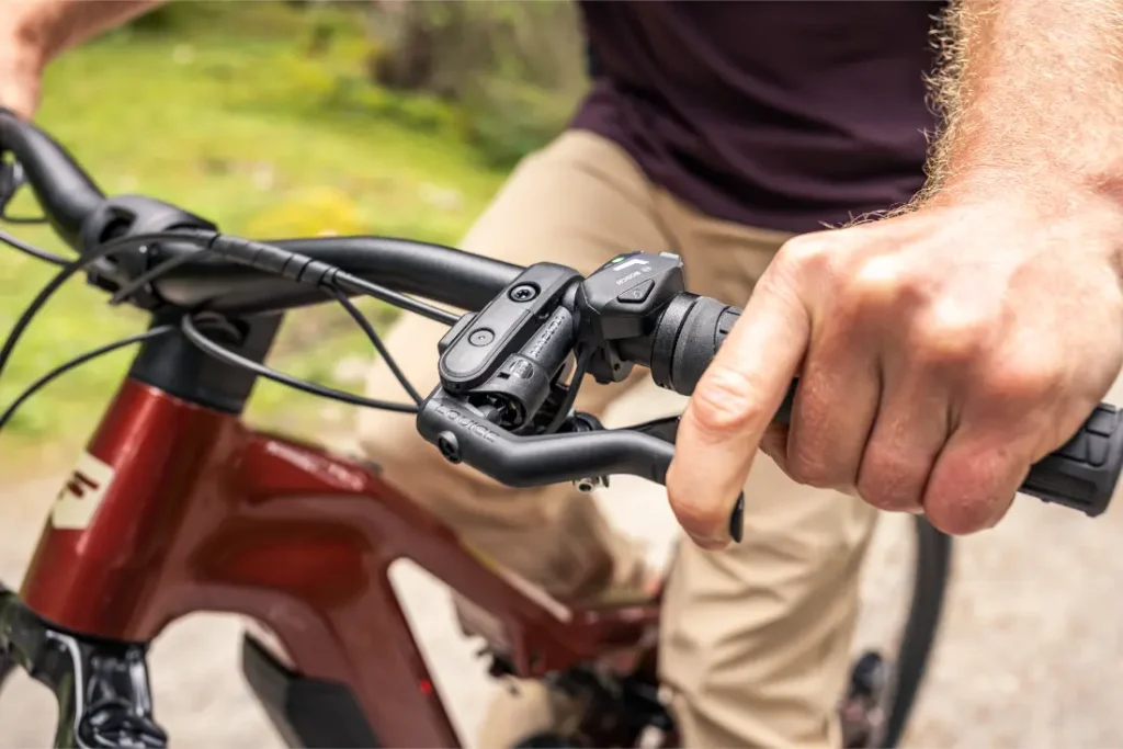 Primer plano de la mano de una persona agarrando el manillar y la palanca de freno de una bicicleta eléctrica de montaña (e-MTB) roja de la marca FOCUS, con vegetación difuminada al fondo.