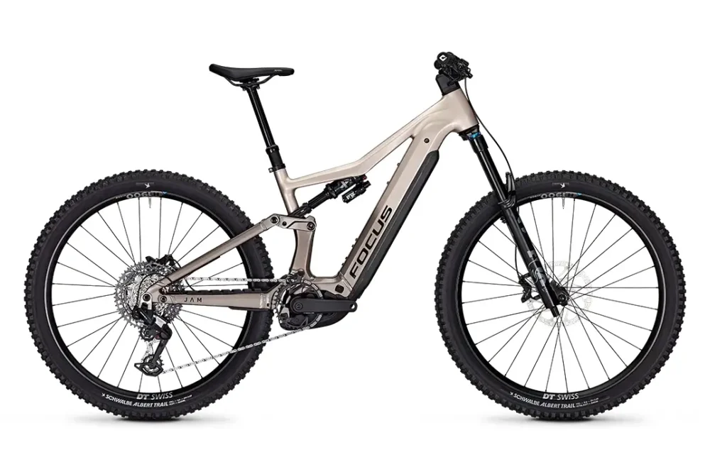 Una bicicleta de montaña eléctrica de doble suspensión FOCUS e-MTB 2027 de color beige con ruedas negras, batería central, frenos de disco y neumáticos anchos con tacos, mostrada de perfil sobre un fondo blanco.