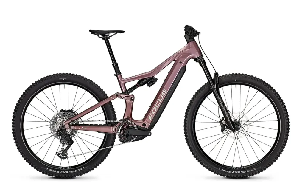 Esta bicicleta eléctrica de montaña FOCUS, de suspensión total y color rosa mate, cuenta con neumáticos de tacos, frenos de disco y un motor de tracción central. La imagen, tomada de perfil sobre un fondo blanco, plasma el espíritu robusto de las series JAM y SAM.