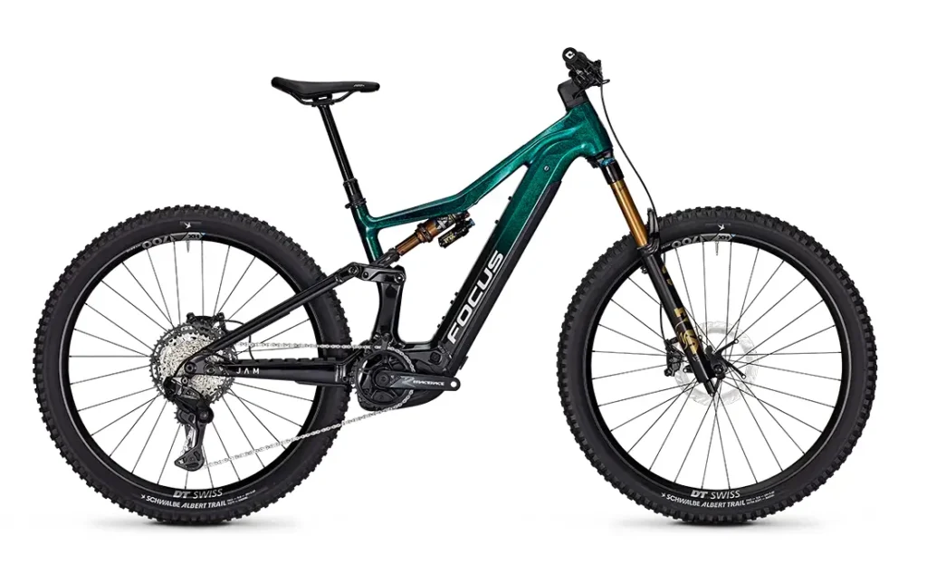 Una bicicleta de montaña Focus e-MTB 2027 de doble suspensión, de color verde y negro, con neumáticos gruesos y con relieve, frenos de disco y un motor eléctrico visible montado en el cuadro.