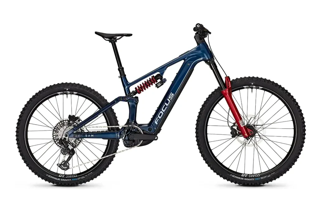 Una bicicleta de montaña eléctrica (e-MTB) FOCUS de doble suspensión, de color azul y rojo, con neumáticos gruesos, frenos de disco y un amortiguador helicoidal visible, fotografiada de perfil sobre un fondo blanco.
