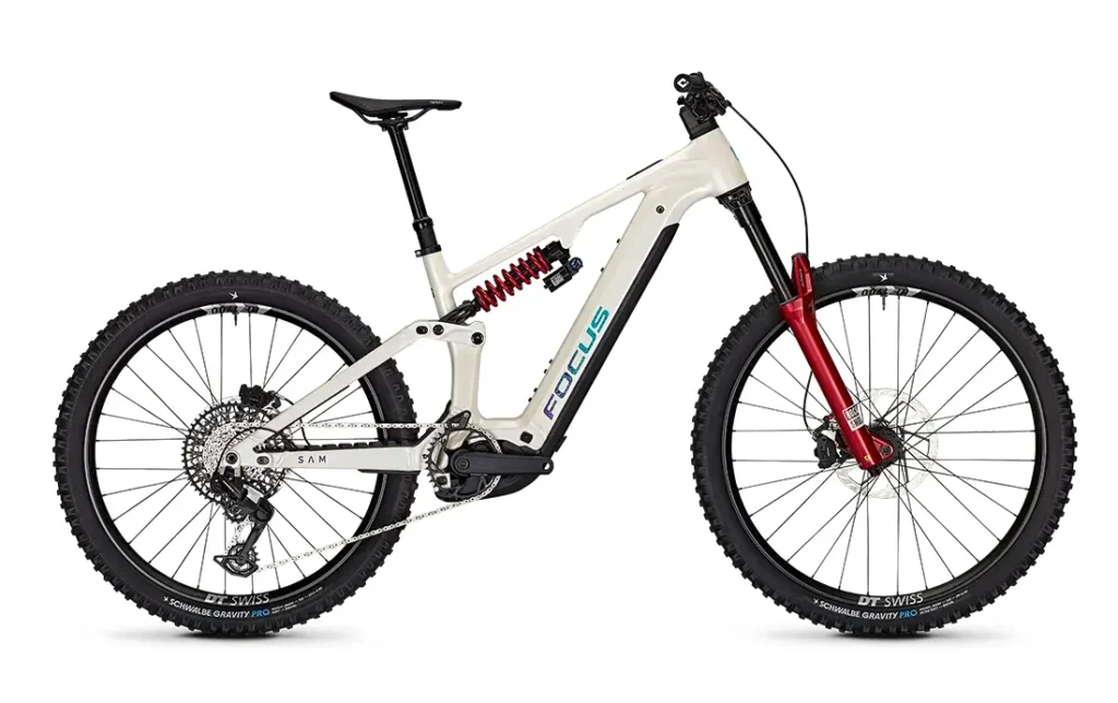 Una bicicleta de montaña eléctrica FOCUS e-MTB de suspensión total, en colores blanco y rojo, con neumáticos de tacos, frenos de disco y el logotipo de Focus en el cuadro; se muestra de perfil sobre un fondo blanco. Ideal para los aficionados a las series JAM o SAM que buscan tanto estilo como rendimiento.