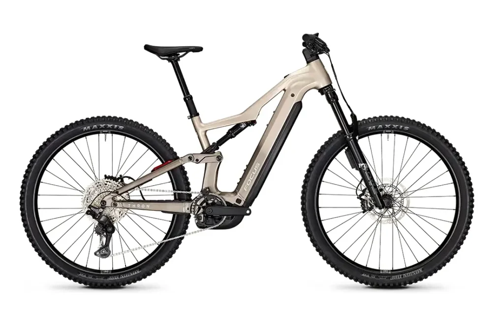 Bicicleta de montaña eléctrica de suspensión total en color beige, con neumáticos anchos, frenos de disco, motor de tracción central y batería visible integrada en el cuadro; forma parte de la innovadora gama Gama FOCUS e-MTB 2027, pensada para ciclistas que buscan estilo y rendimiento.