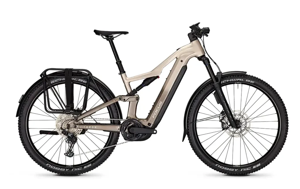 Una bicicleta eléctrica FOCUS e-MTB de color beige con motor central, frenos de disco, portaequipajes trasero, horquilla de suspensión delantera y neumáticos grandes con banda de rodadura.