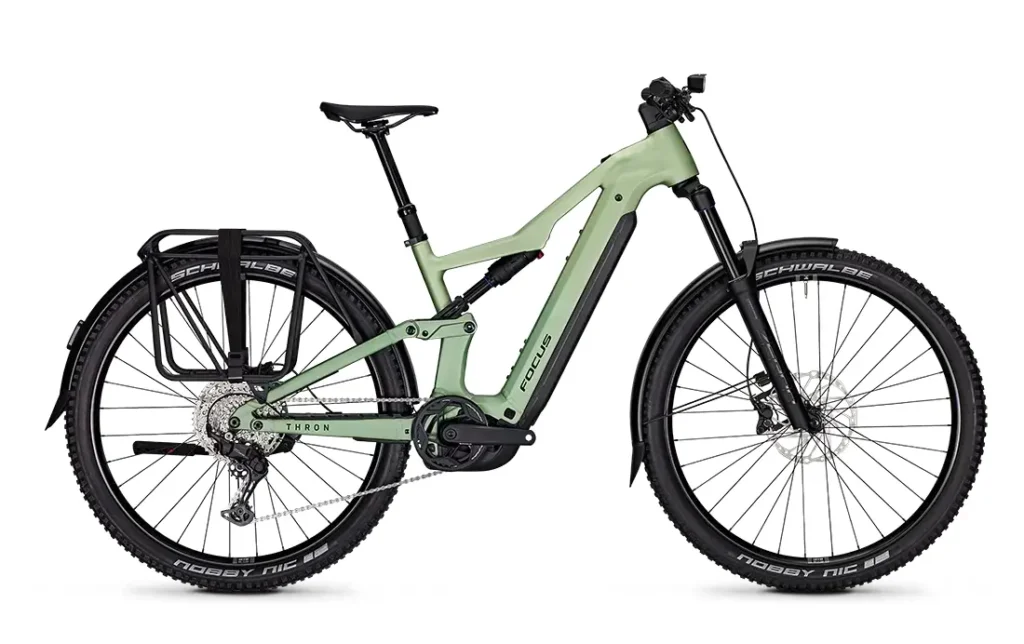 Bicicleta de montaña eléctrica FOCUS e-MTB 2027 de color verde claro, con batería negra, portaequipajes trasero, guardabarros, horquilla de suspensión y neumáticos Schwalbe de gran tamaño; se muestra de perfil sobre un fondo blanco.