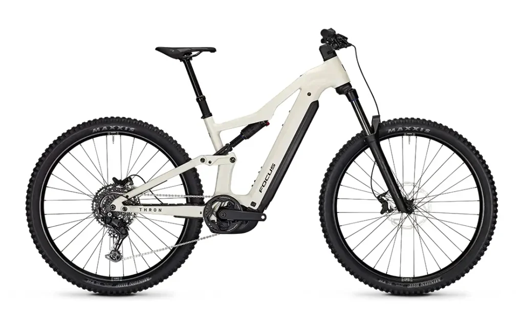 Una bicicleta eléctrica de montaña (e-MTB) FOCUS de suspensión total, de color blanco, con ruedas negras, frenos de disco y un motor central visible; este elegante modelo JAM está diseñado para ciclistas que buscan tanto rendimiento como estilo en los senderos.