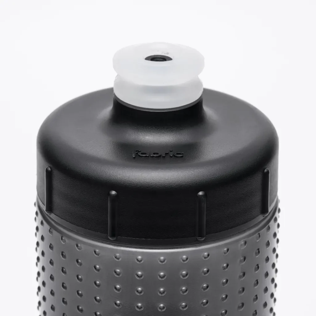Primer plano de la parte superior de una botella de agua negra con textura y una boquilla translúcida que se levanta para beber. La palabra «fabric» aparece grabada en relieve en la botella, justo debajo del tapón, lo que resalta el elegante diseño del bidón antimicrobiano Fabric Gripper V2, que ofrece protección permanente.