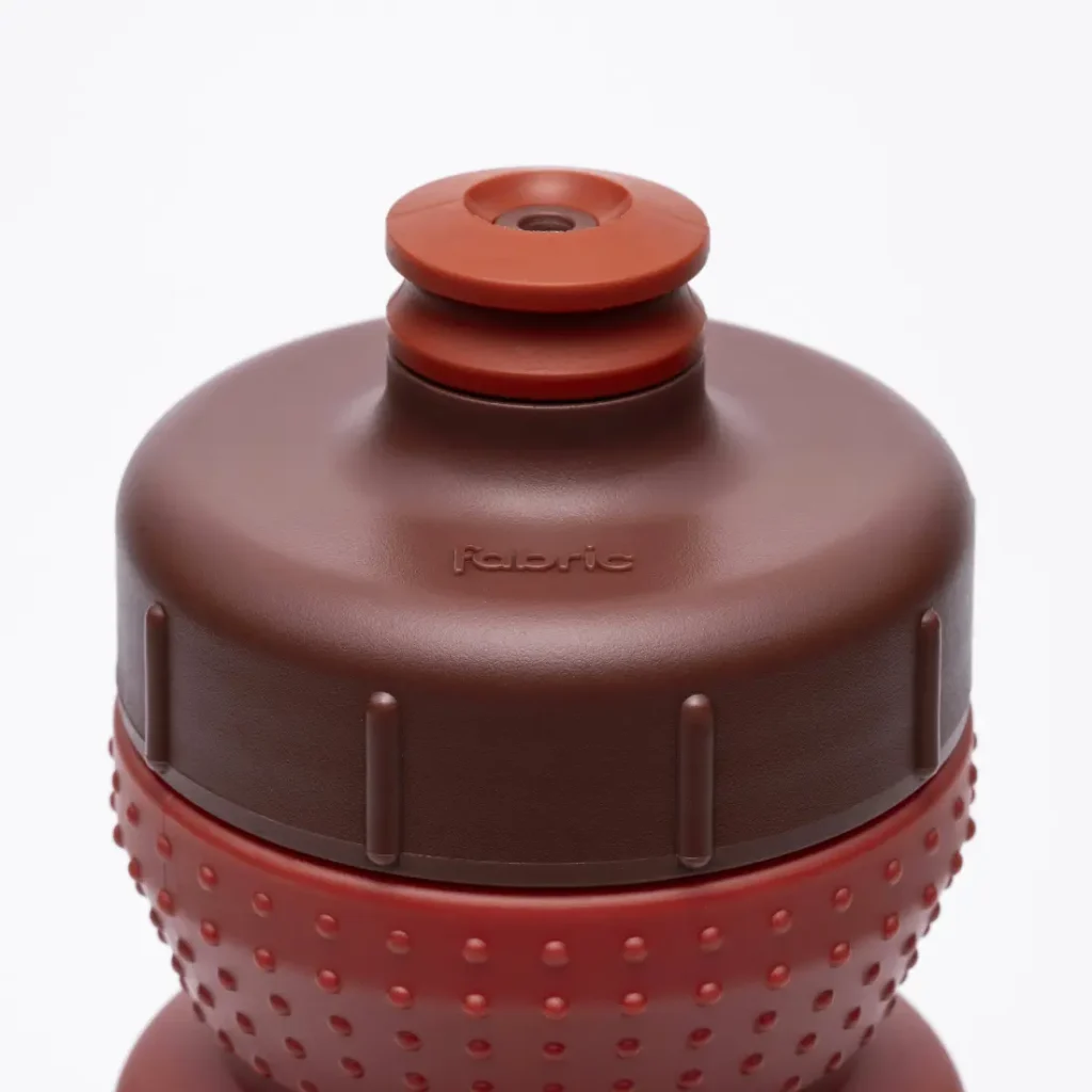 Primer plano de una botella de agua de plástico «Fabric Gripper V2» de color marrón y rojo, con un agarre texturizado y la palabra «fabric» grabada en relieve en el tapón. Esta botella antimicrobiana ofrece protección permanente, lo que la hace ideal para la hidratación diaria.