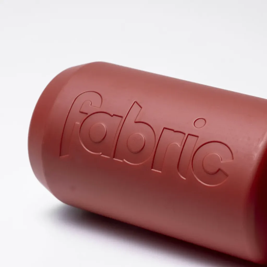 Un primer plano de un recipiente cilíndrico rojo, el Fabric Gripper V2, con la palabra «fabric» grabada en relieve en uno de sus laterales, sobre un fondo blanco liso. Este bidón antimicrobiano está diseñado para ofrecer una protección permanente.