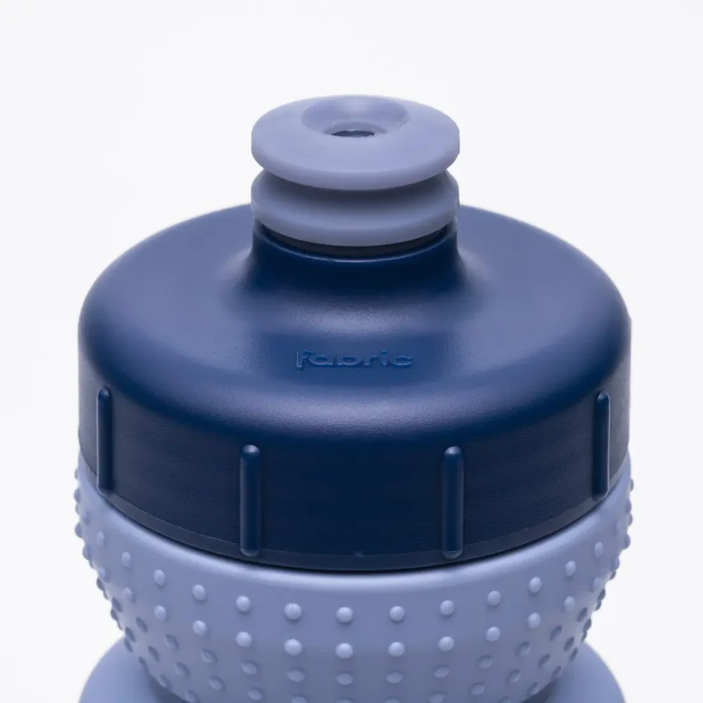 Primer plano de una botella deportiva azul y gris con un agarre texturizado y una boquilla de tipo «push-pull». La palabra «Fabric» aparece grabada en relieve en la tapa, lo que pone de relieve el avanzado diseño Fabric Gripper V2 para un manejo cómodo. Esta botella antimicrobiana también ofrece protección permanente para ayudarte a mantener tu hidratación limpia y segura.