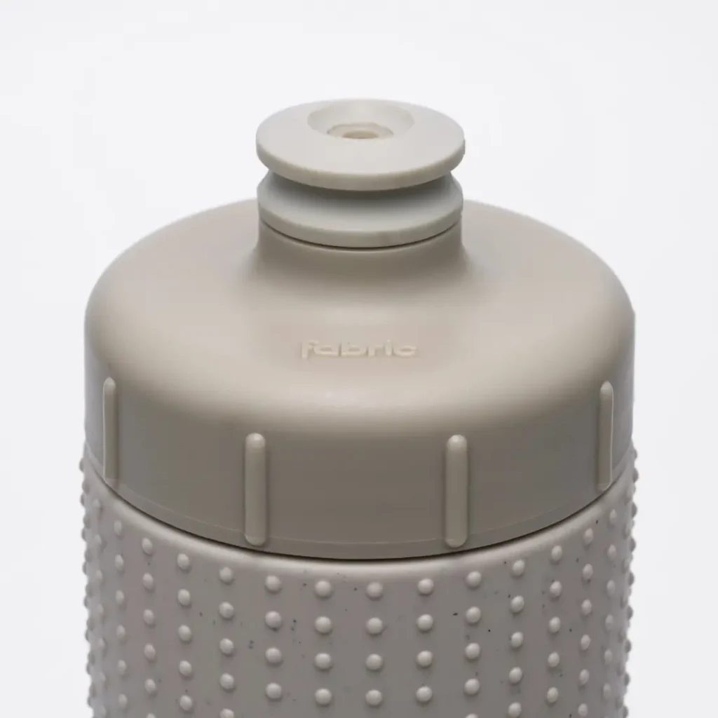 Primer plano de una tapa para botella de agua «Fabric Gripper V2» de color gris claro, con una boquilla de tipo «push-pull» y una textura de puntos en relieve en la parte superior de la botella; la palabra «fabric» aparece grabada en relieve en la tapa, lo que destaca sus propiedades antimicrobianas para una protección permanente.