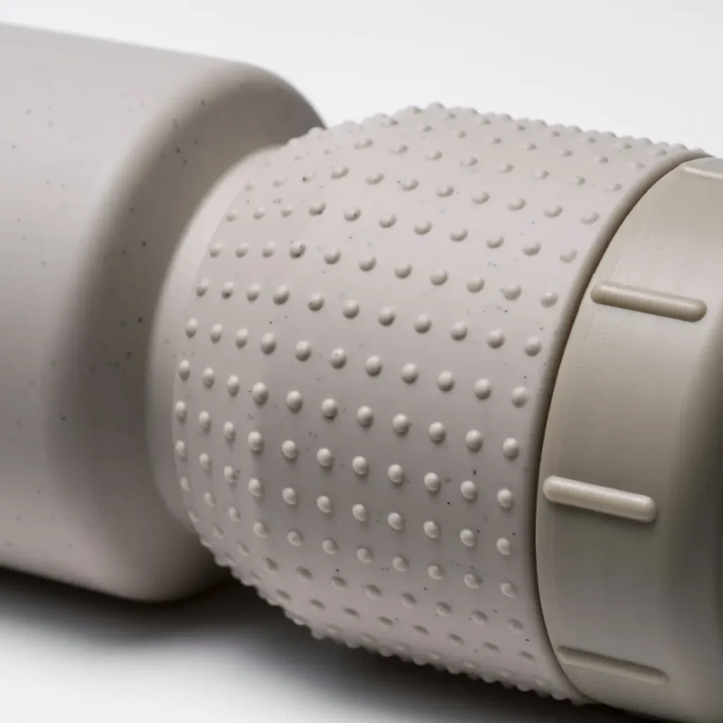 Primer plano de un objeto cilíndrico de color gris claro y con textura —concretamente, el bidón antimicrobiano «Fabric Gripper V2»— con una zona de agarre cubierta de pequeñas protuberancias y un tapón liso en el extremo.