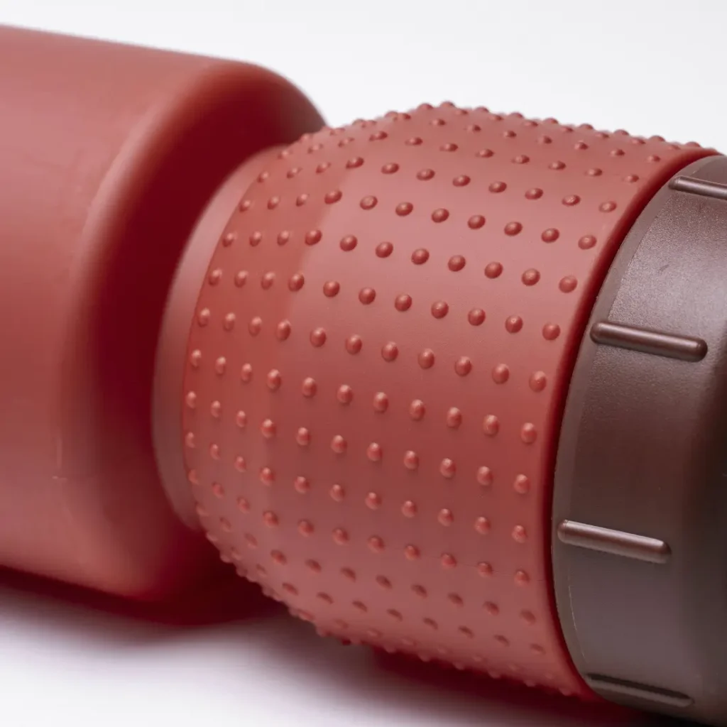 Primer plano de un agarre de goma rojo con textura —conocido como «Fabric Gripper V2»— en un bidón cilíndrico con tapón de color marrón oscuro, sobre un fondo blanco. Este diseño incorpora protección antimicrobiana para una mayor higiene.