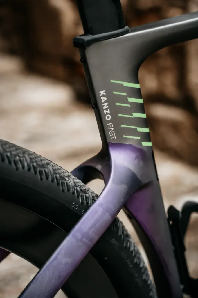 Primer plano del cuadro y la rueda trasera de una bicicleta Ridley Kanzo Fast 2.0, con el logotipo «KANZO FAST» y líneas gráficas verdes sobre un cuadro negro mate y morado, que resaltan su diseño aerodinámico y su compatibilidad con neumáticos de 58 mm para un rendimiento óptimo.