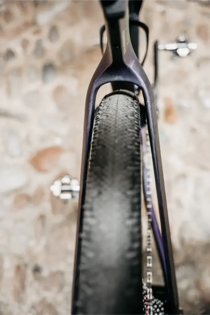 Primer plano del neumático trasero y el cuadro de una Ridley Kanzo Fast 2.0, con un muro de piedra al fondo, en el que destacan el diseño aerodinámico, el marcado dibujo del neumático y la estructura robusta, equipada con neumáticos de 58 mm.