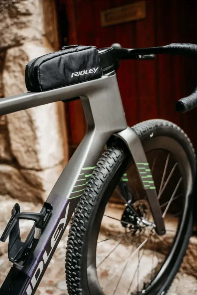 Primer plano de una bicicleta Ridley Kanzo Fast 2.0 con una pequeña bolsa negra de Ridley sujeta al manillar, que presenta detalles de diseño aerodinámico y neumáticos anchos de 58 mm, aparcada junto a un muro de piedra y una puerta de madera.