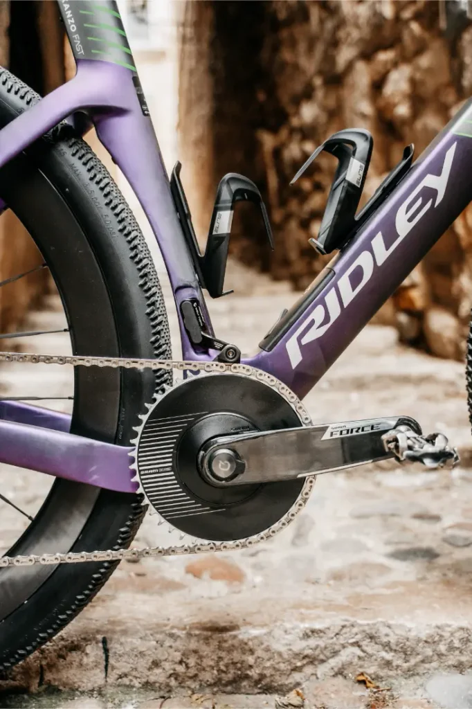 Primer plano de la transmisión de una bicicleta Ridley Kanzo Fast 2.0, en el que se aprecian el juego de bielas, la cadena, los pedales y parte del cuadro aerodinámico con dos portabidones vacíos. La bicicleta está aparcada sobre una superficie de piedra, lista para rodar con sus robustos neumáticos de 58 mm.