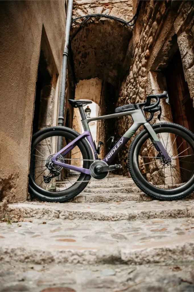 Una moderna bicicleta de carretera, la Ridley Kanzo Fast 2.0, con un cuadro morado y gris y características aerodinámicas, está aparcada en unos escalones de piedra entre antiguos y estrechos edificios de piedra en un callejón.