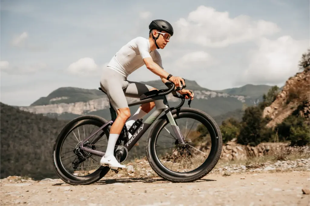 Un ciclista, con casco y equipación de ciclismo, circula en una bicicleta de gravel Ridley Kanzo Fast 2.0 con neumáticos de 58 mm por un sendero rocoso, con montañas y árboles al fondo.