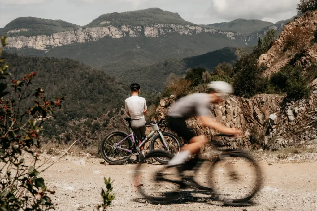 Dos ciclistas en una ruta de montaña; uno está de pie junto a una Ridley Kanzo Fast 2.0 —con su cuadro aerodinámico y sus neumáticos de 58 mm, preparados para cualquier terreno—, de cara a unos acantilados lejanos, mientras que el otro pasa en bicicleta por el primer plano, difuminado por el movimiento.