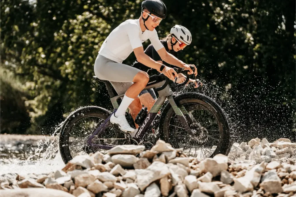 Dos ciclistas, con casco y gafas de sol, pedalean en unas bicicletas de gravel Ridley Kanzo Fast 2.0, equipadas con manillares aerodinámicos y robustos neumáticos de 58 mm, a través de un arroyo poco profundo y rocoso, salpicando agua mientras pedalean. Al fondo se ven árboles y vegetación.