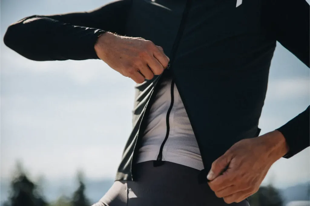 Una persona se abrocha una chaqueta negra ASSOS EQUIPE sobre una camisa blanca al aire libre; se le ve parte del torso y las manos sobre un fondo difuminado, lo que destaca la protección ultraligera y las innovadoras características del ThermoBooster.