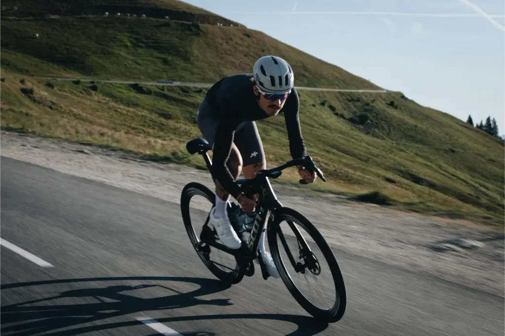 Una persona que lleva casco y equipación de ciclismo, incluido el ASSOS EQUIPE ThermoBooster para una protección ultraligera en condiciones de entretiempo, desciende en bicicleta de carretera por una carretera asfaltada junto a colinas cubiertas de hierba bajo un cielo despejado.