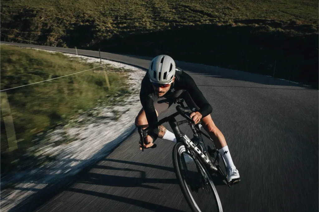 Un ciclista que lleva casco y gafas de sol se inclina al tomar una curva en una carretera asfaltada, con colinas cubiertas de hierba al fondo, confiando plenamente en el ASSOS EQUIPE ThermoBooster para disfrutar de una protección ultraligera durante esos días impredecibles de entretiempo.