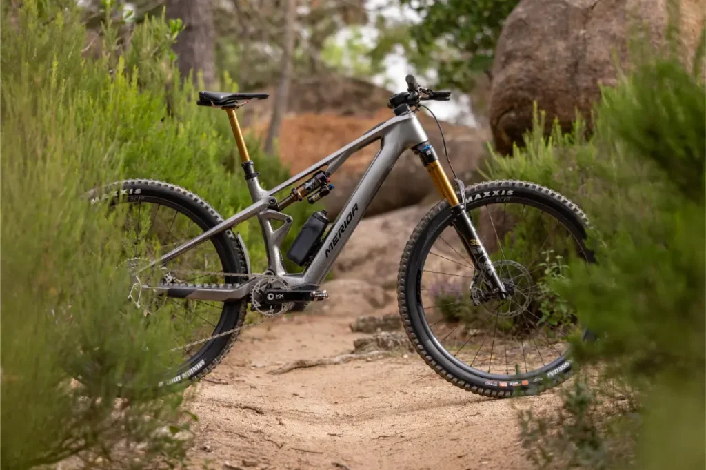 Una bicicleta de montaña gris, con suspensión total y neumáticos gruesos, se encuentra en un sendero de tierra rodeado de matorrales verdes y rocas, en una zona boscosa.